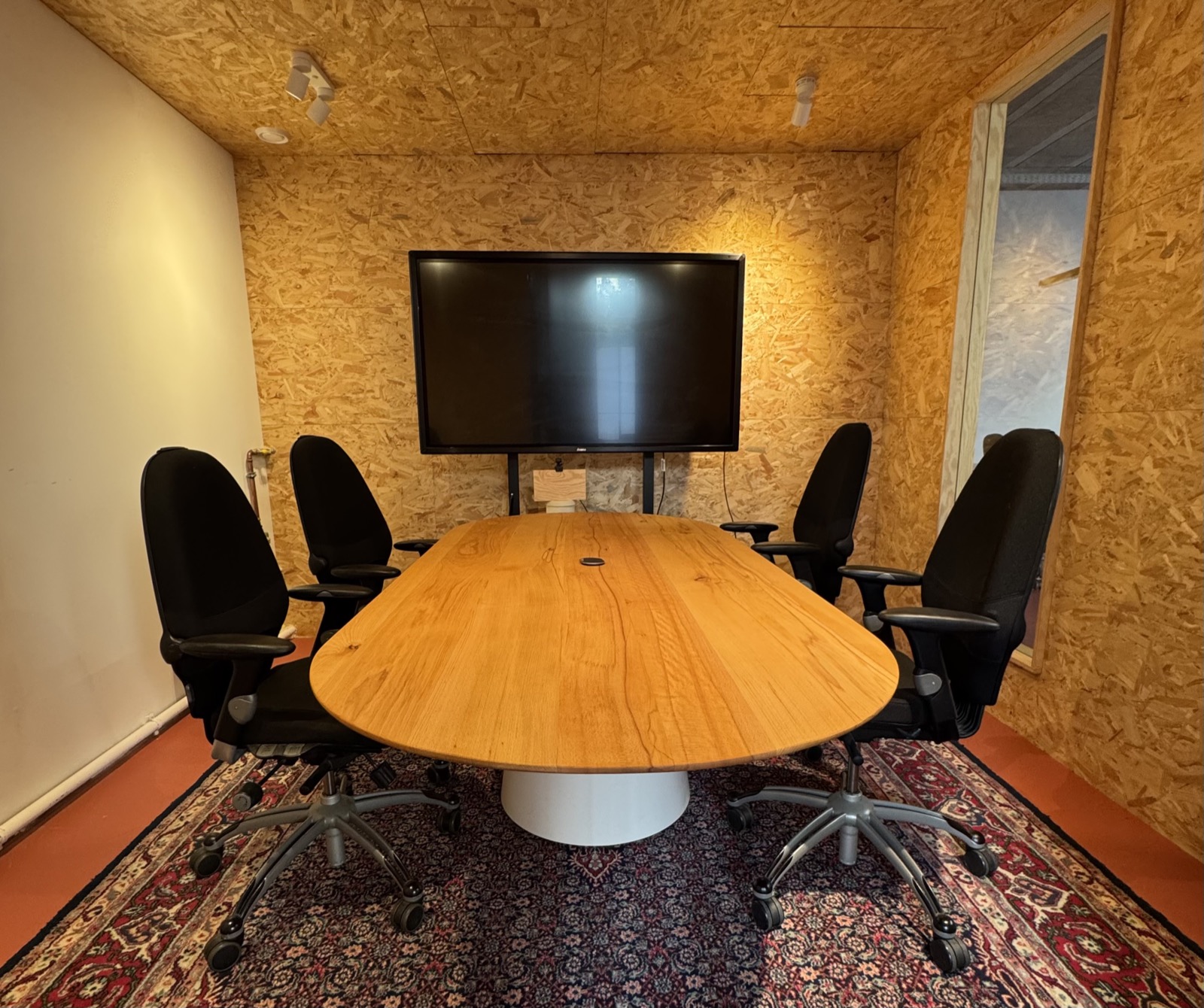The meeting room, lined in OSB, with an oval wooden table and a screen.