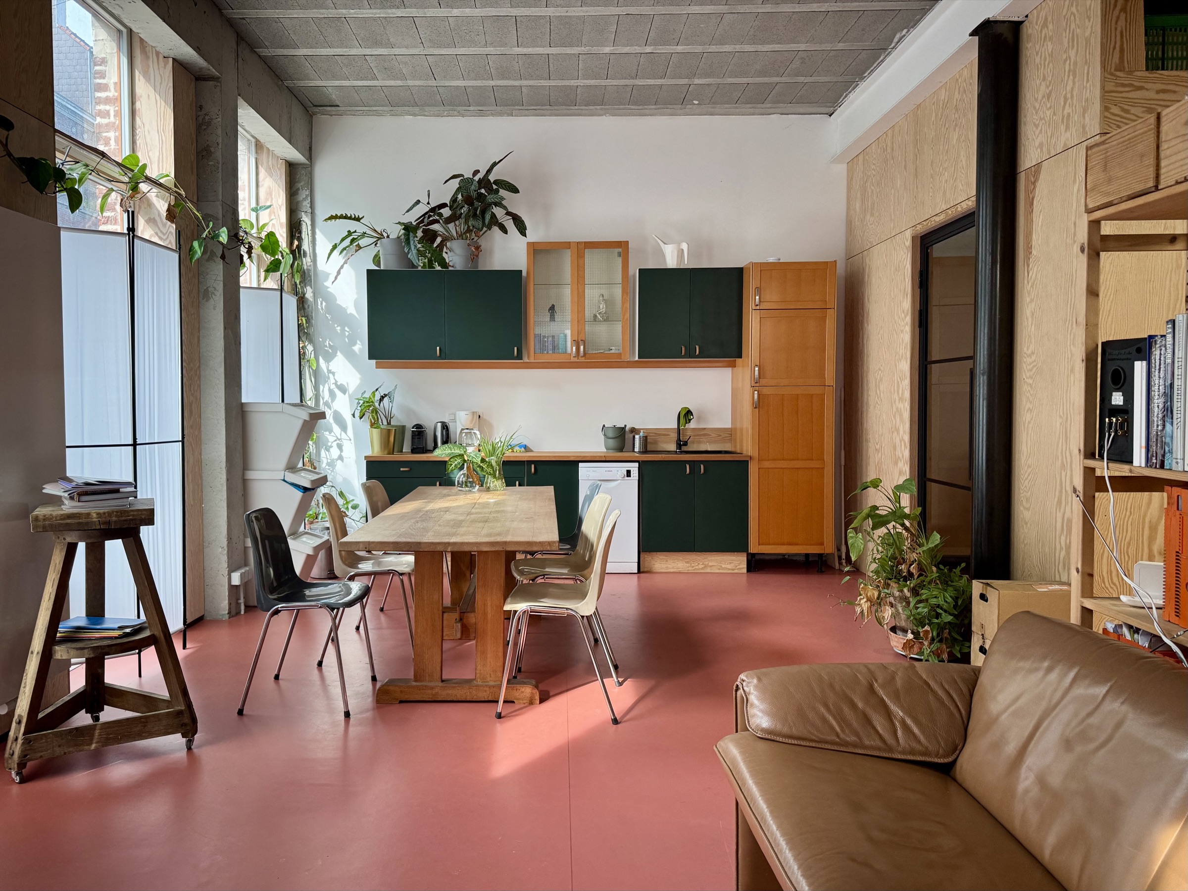 The common room: a long wooden table, green kitchen units and a terracotta floor under a concrete workshop ceiling.